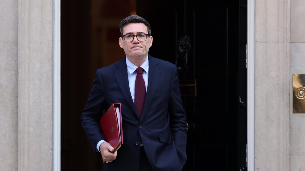 The image shows Andy Burnham walking out of 10 Downing Street. He is wearing a navy suit, white shirt, burgundy tie and glasses. He is holding a red file.