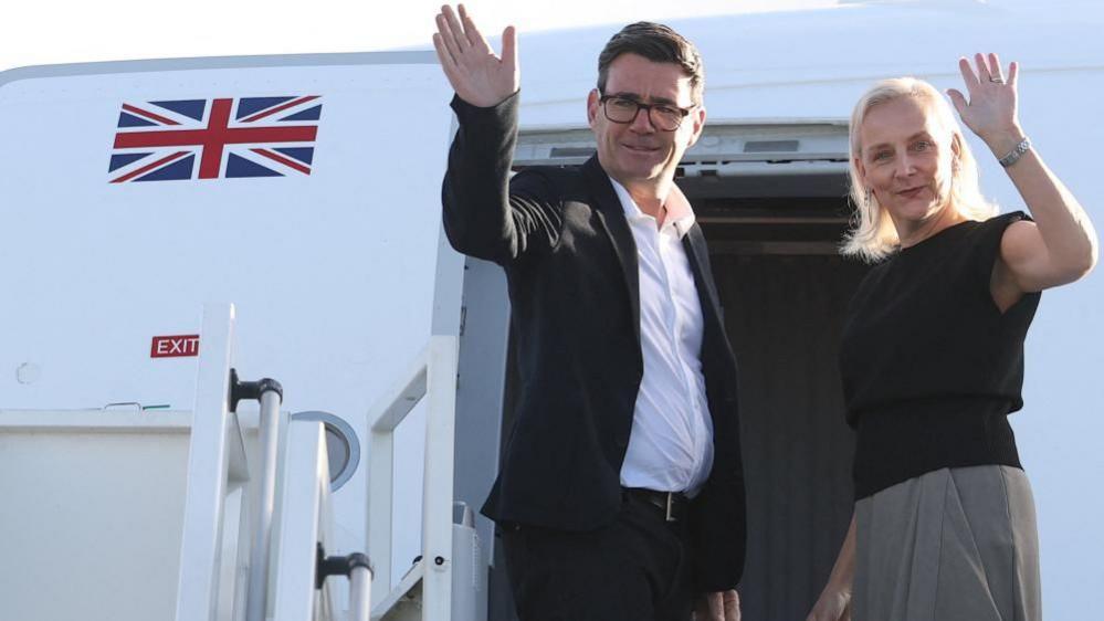 Andy Burnham and Marie-France Van Heel wave as they stand at the top of the steps to an aircraft. Burnham is wearing a black jacket and trousers, a white shirt and glasses. Van Heel is wearing a black top, a grey skirt and has a watch on her left wrist. The aircraft is white and the Union flag also features as part of its livery.