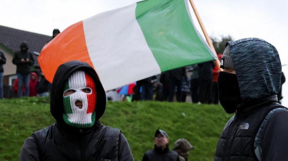 A person wears an Irish flag themed mask with another person next to them wearing a beanie and ski mask with protesters and the Irish flag behind them