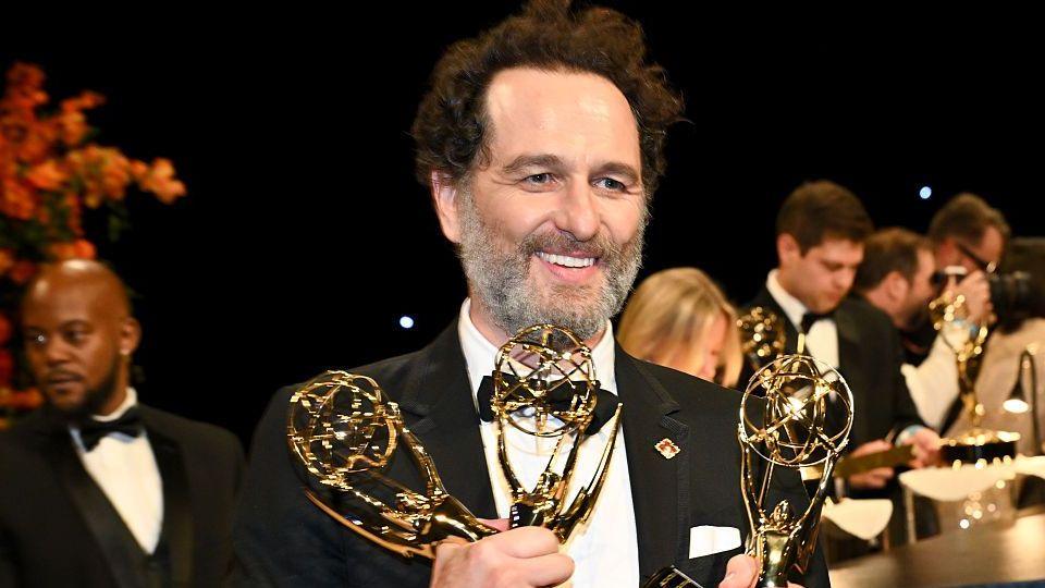 Matthew Rhys is photographed attending the 78th Emmy Awards. He is wearing a black suit jacket, black waist coat and black bow tie. He has dark hair and a grey beard. He is smiling whilst holding three Emmy awards. He is looking away from the camera.