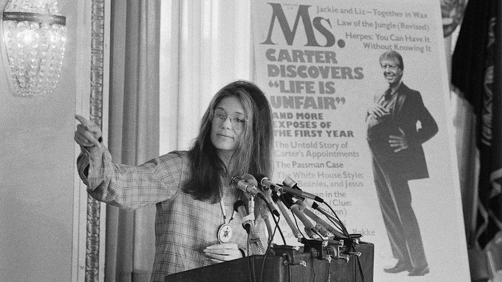Gloria Steinem standing in front of a cover of Ms. magazine