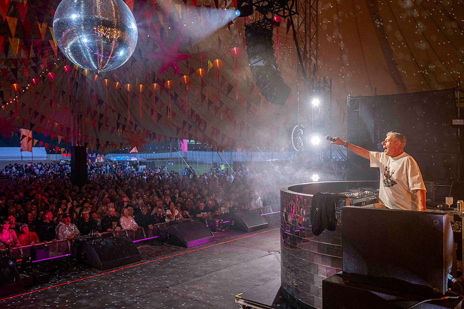 Gary Davies DJing on stage. His arm, holding a microphone, is outstretched towards a large crowd.