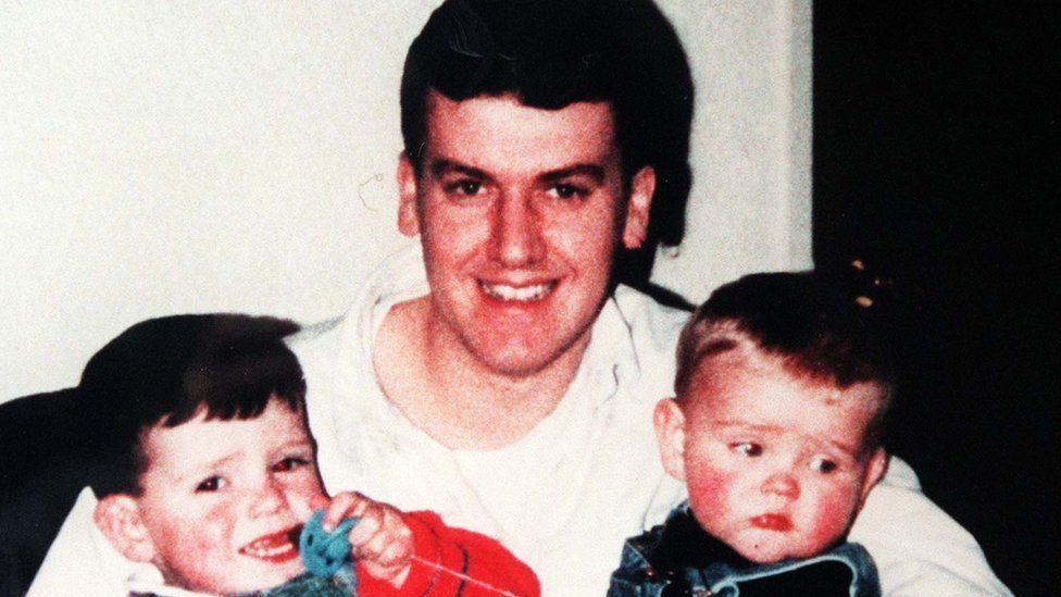 A man in a whilte shirt with two toddlers on his knee.