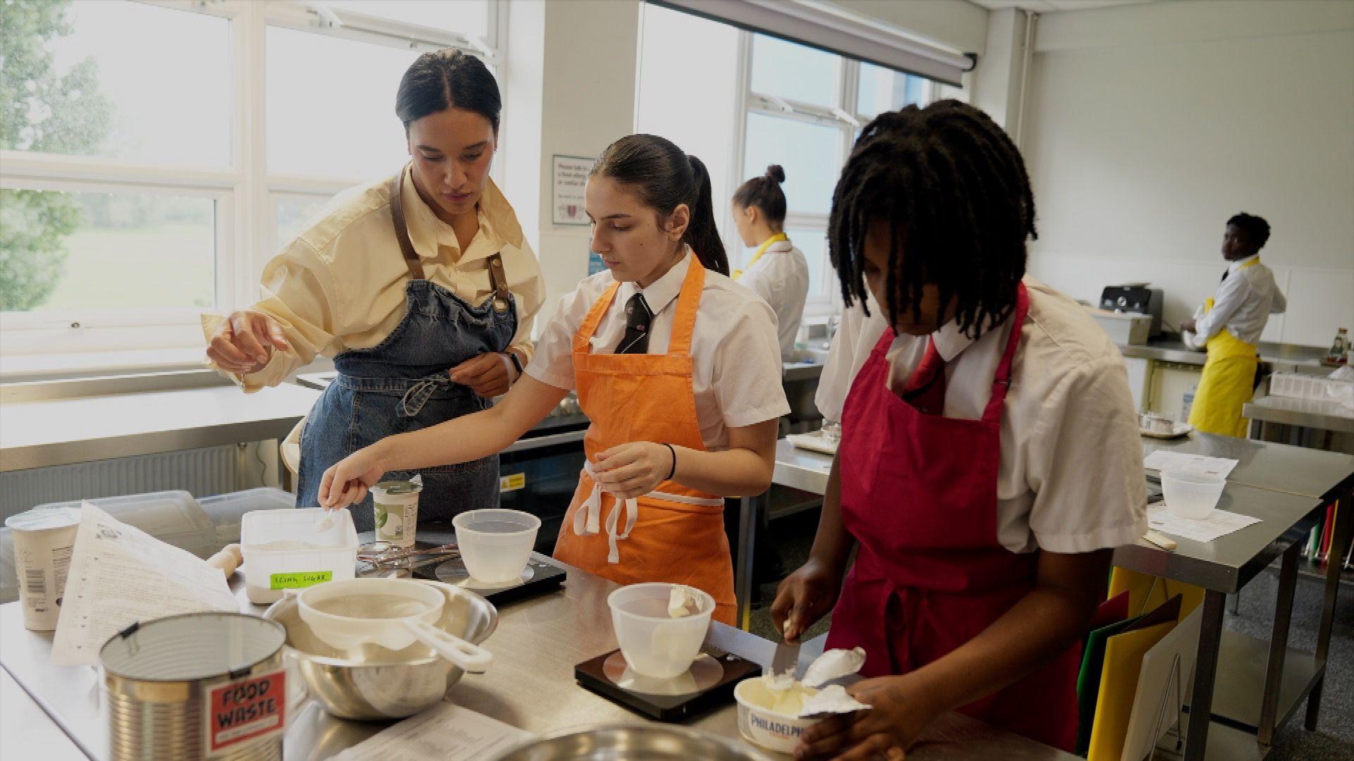 Two teenagers at a classroom bench, wearing aprons., They are in a catering class and are adding cream cheese to bowls, and are being supervised by their female teacher, also in an apron, who is standing to the left of them.