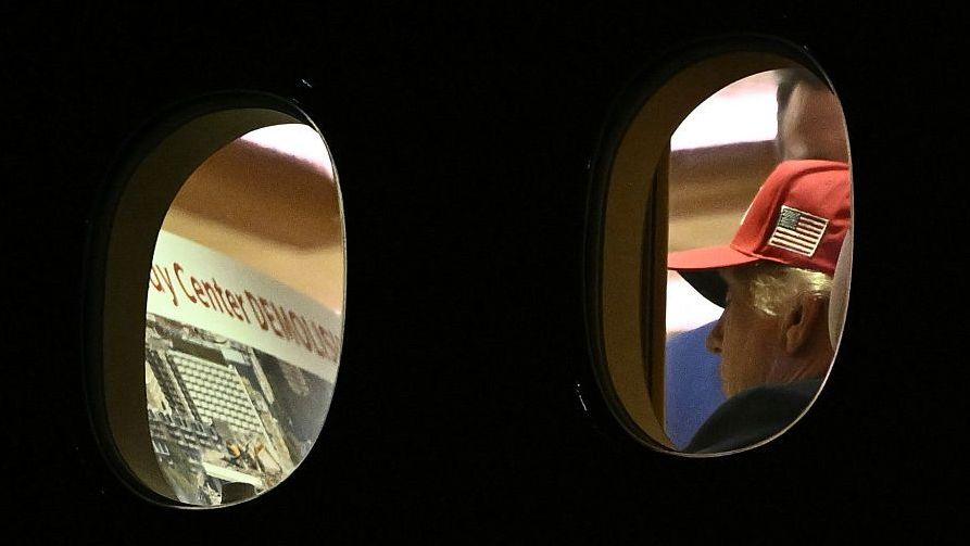 President Donald Trump photographed through the windows of Air Force One looking at a poster about the Kennedy Center. he is wearing a red cap