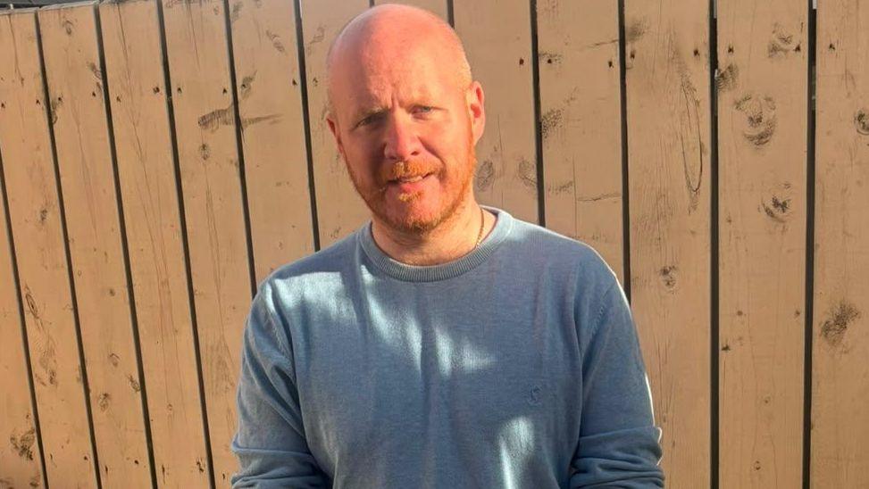 A bald man with ginger stubble looking at the camera, standing in the sunshine in front of a wooden fence.