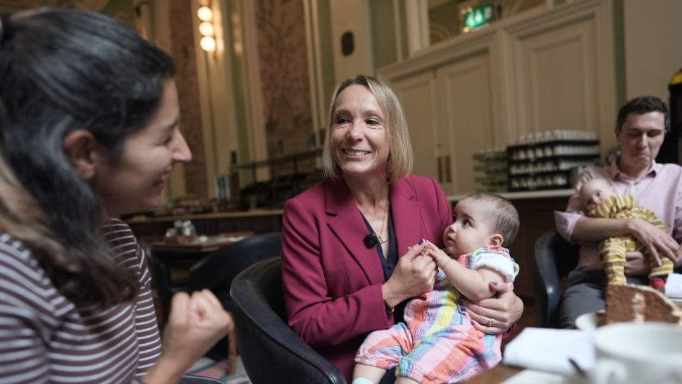 Helen Morgan is in the centre of the picture, smiling and holding a baby on her lap. Morgan has a blonde bob and is wearing a berry-coloured jacket over a navy top. She is looking at a woman in a stripy top on the left and a man holding another baby is on the right.