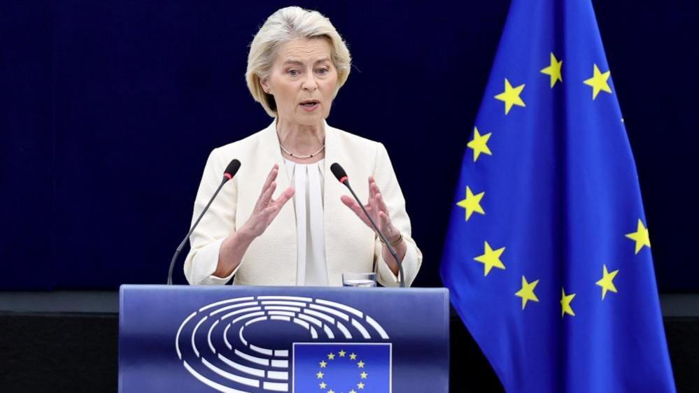 Ursula von der Leyen, who has a grey/blonde short haircut, wears a cream blazer and blouse as she speaks at a podium, with the EU flag in the background