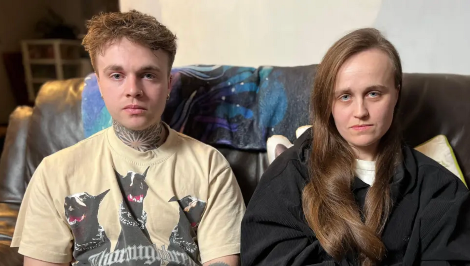 Two young people sit on a sofa is a man with a neck tattoo, short brown hair and wearing a beige T-shirt with a black pattern on it. The other is a young woman wearing a black top. She has thick, long brown hair.