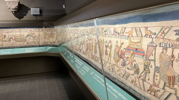 The Bayeux Tapestry depicting the Norman conquest in 1066 is pictured behind a glass case along a grey wall.