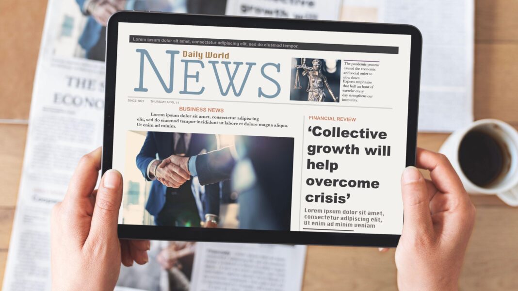 A person's hands holding a tablet with a mock-up of a news website over a table with a newspaper and cup of coffee
