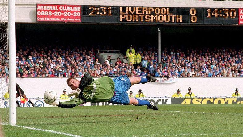 Knight diving full length to make a save with his outstretched right hand against Liverpool, wearing a green goalkeeping jersey and blue shorts and socks
