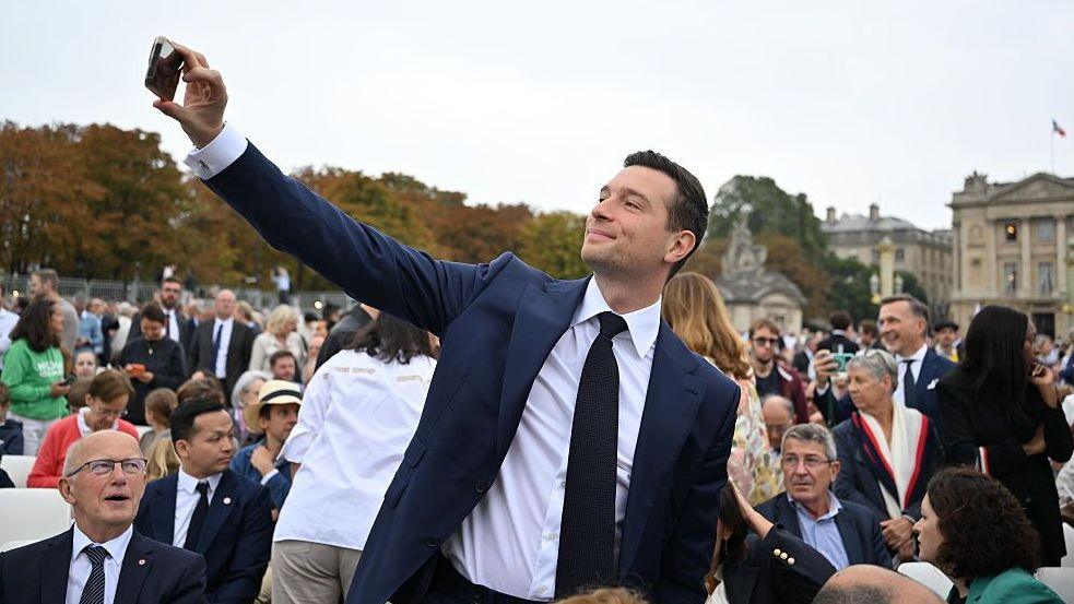 Jordan Bardella, wearing a suit and tie, takes a selfie in a crowd during the Pope's solemn outdoor mass at Place de la Concorde on the second day of his visit to Paris