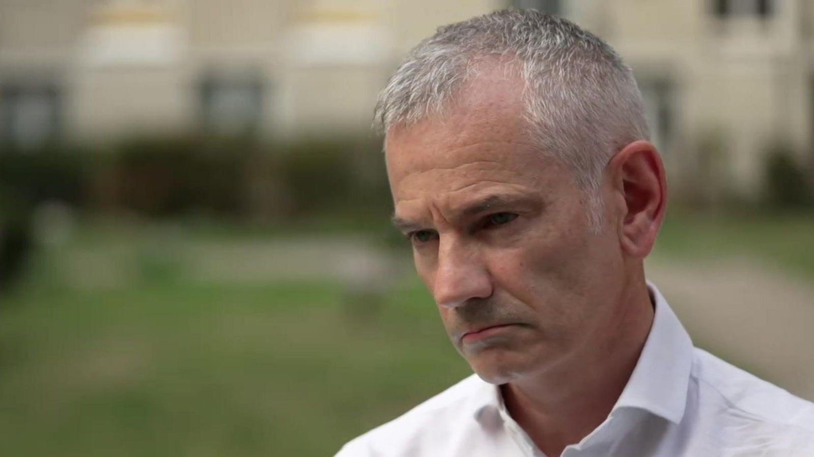 A middle-aged man with white hair and wearing an open necked white shirt being interviewed in a park.