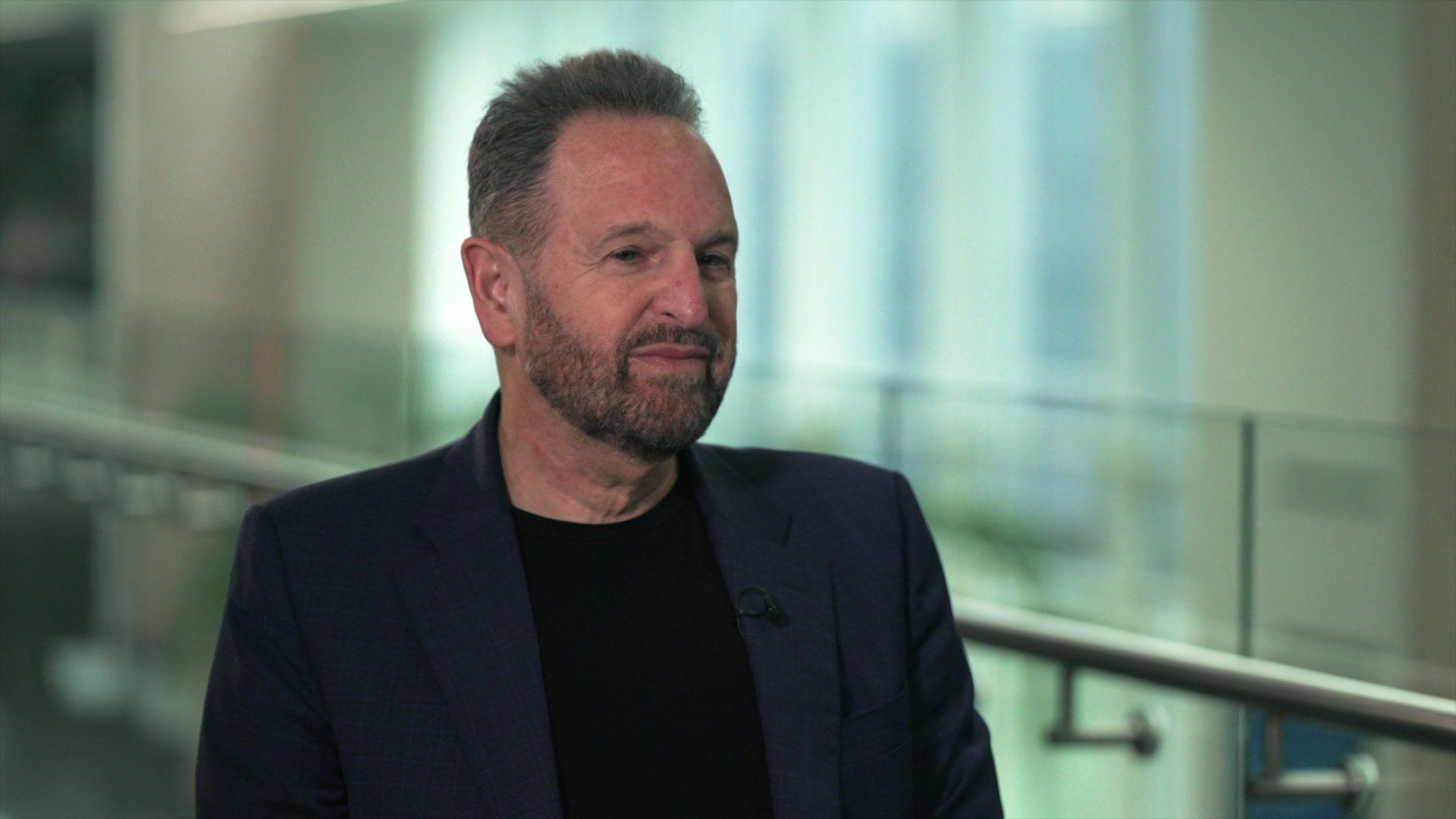 Rene Haas sitting being interviews, wearing a dark blue blazer over a black T-shirt