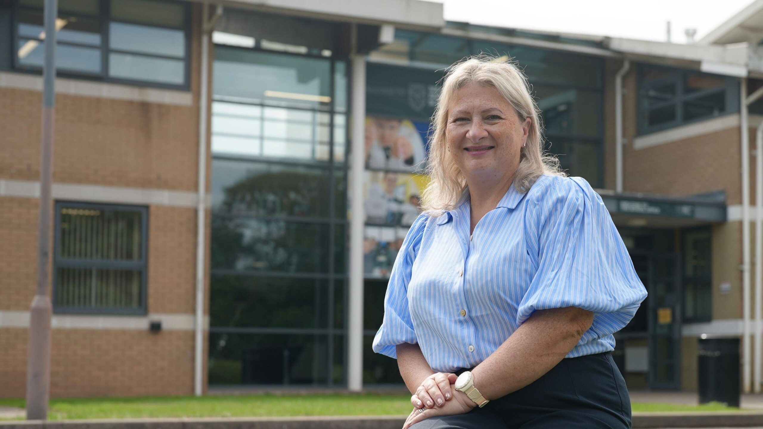 Bernadette Pettman is seen seated on a stone bench outside the modern school building, wearing a light blue striped blouse and dark trousers. Large glass windows and brick walls form the background, with a parked car visible to one side.