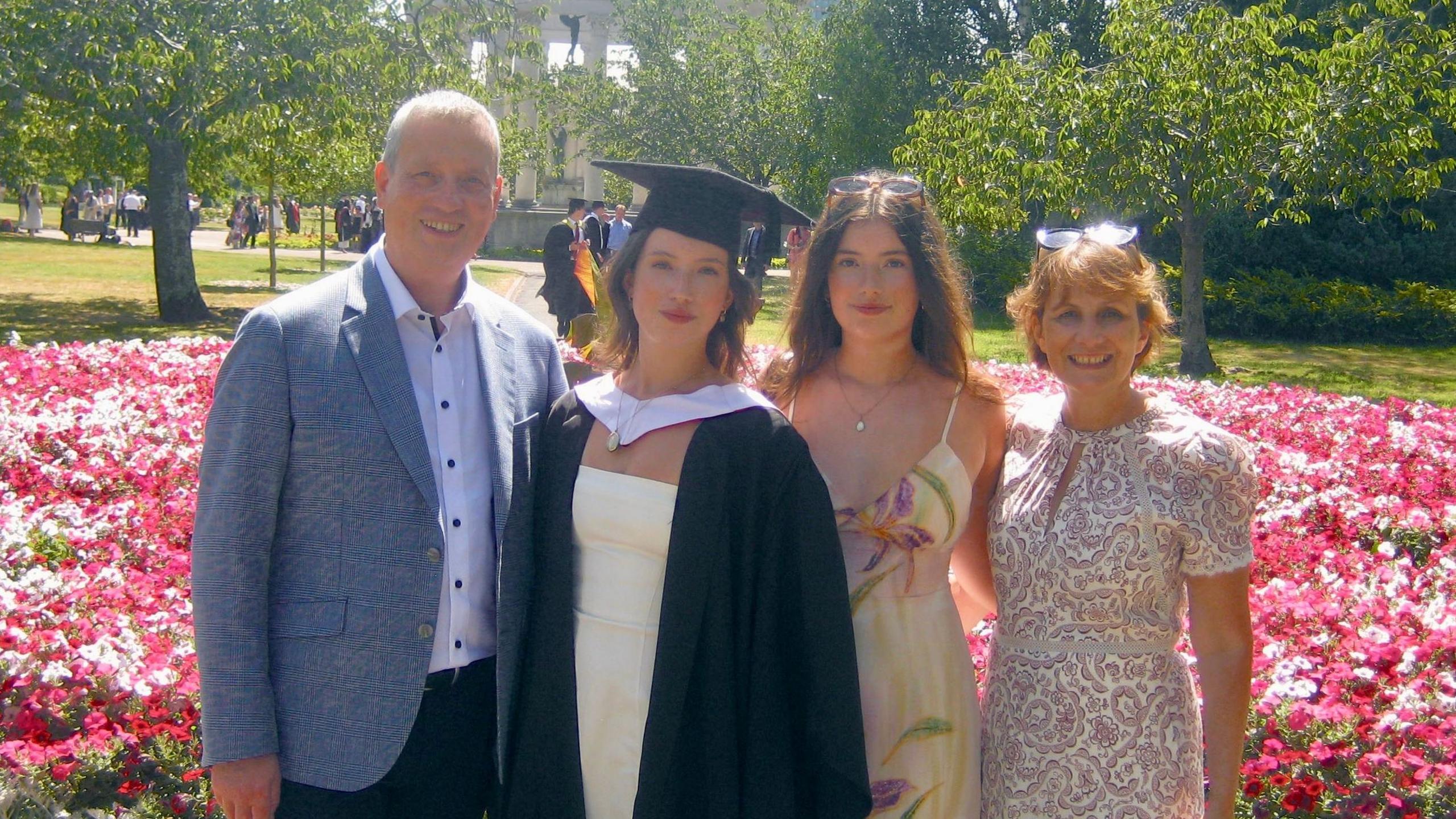 A family of four stood in a row with a pink flowerbed and trees behind them, all smiling and looking at the camera. Left to right: a middle-aged man with grey hair wearing a white shirt and grey suit jacket; a young woman with long dark hair wearing a white dress and black graduaiton cap and gown; a young woman with long dark hair wearing a yellow floral-patterned dress and sunglasses on her head; and a middle-aged woman wiht short brown hair, wearing a pink and white paisley-pattern dress and sunglasses on her head.