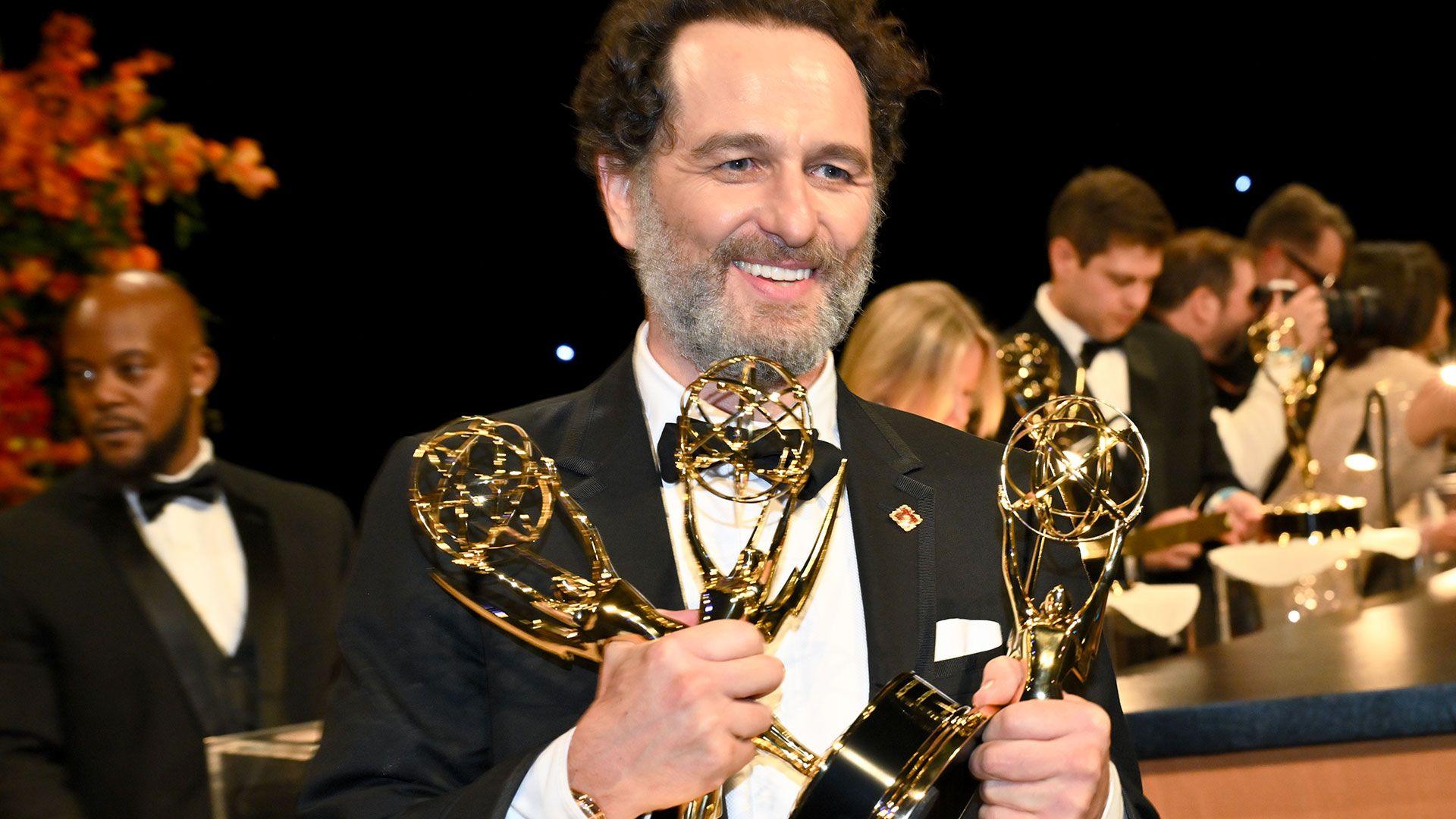Matthew Rhys is photographed attending the 78th Emmy Awards. He is wearing a black suit jacket, black waist coat and black bow tie. He has dark hair and a grey beard. He is smiling whilst holding three Emmy awards. He is looking away from the camera.