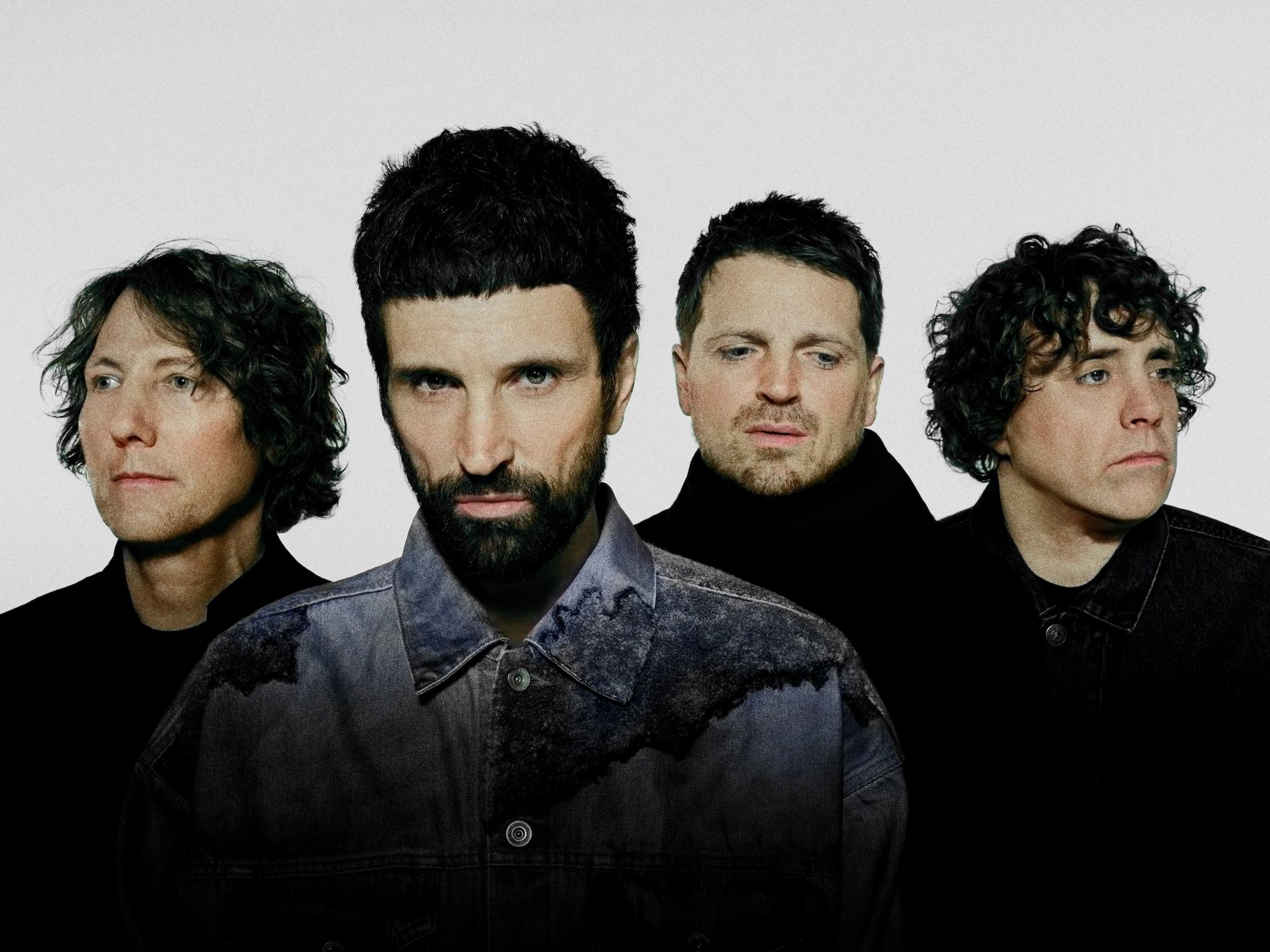 Studio portrait of Kasabian. Three band members stand at the back wearing black, while Serge wears a denim shirt.