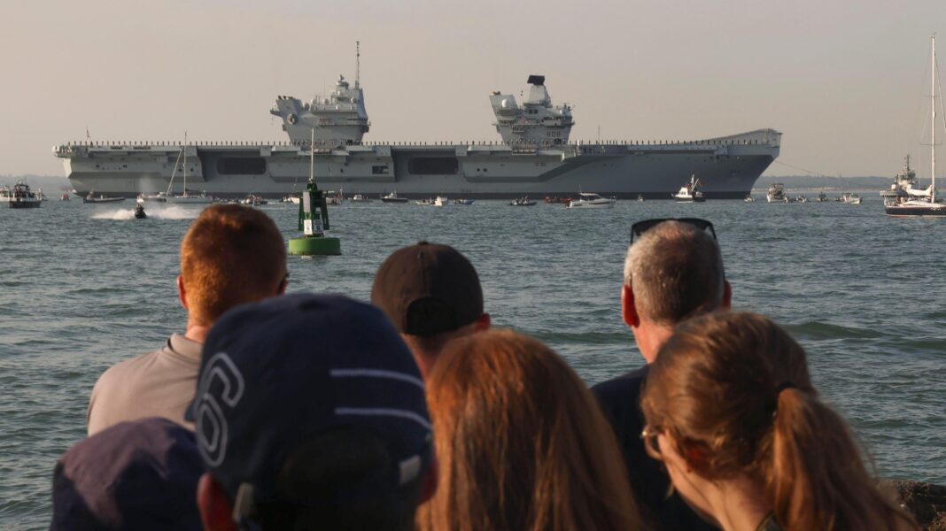 Royal Navy aircraft carrier departs for training A group of people out of focus at the front with the queen elizabeth on the water in the background. The sky is orange.