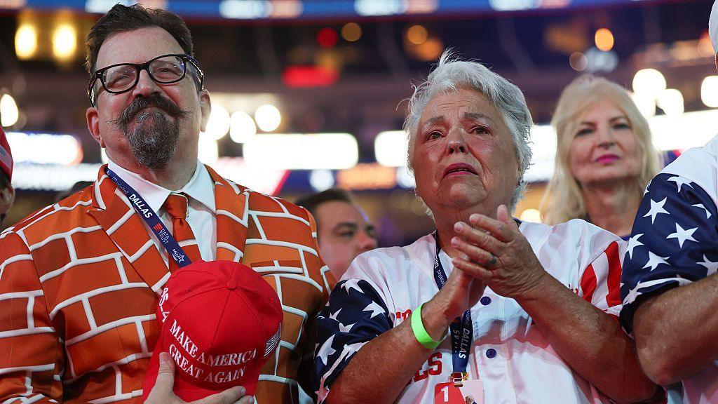 A man in a brick wall suit stands during the convention. He is holding a Make America Great Again hat. Beside him, a woman stands clapping