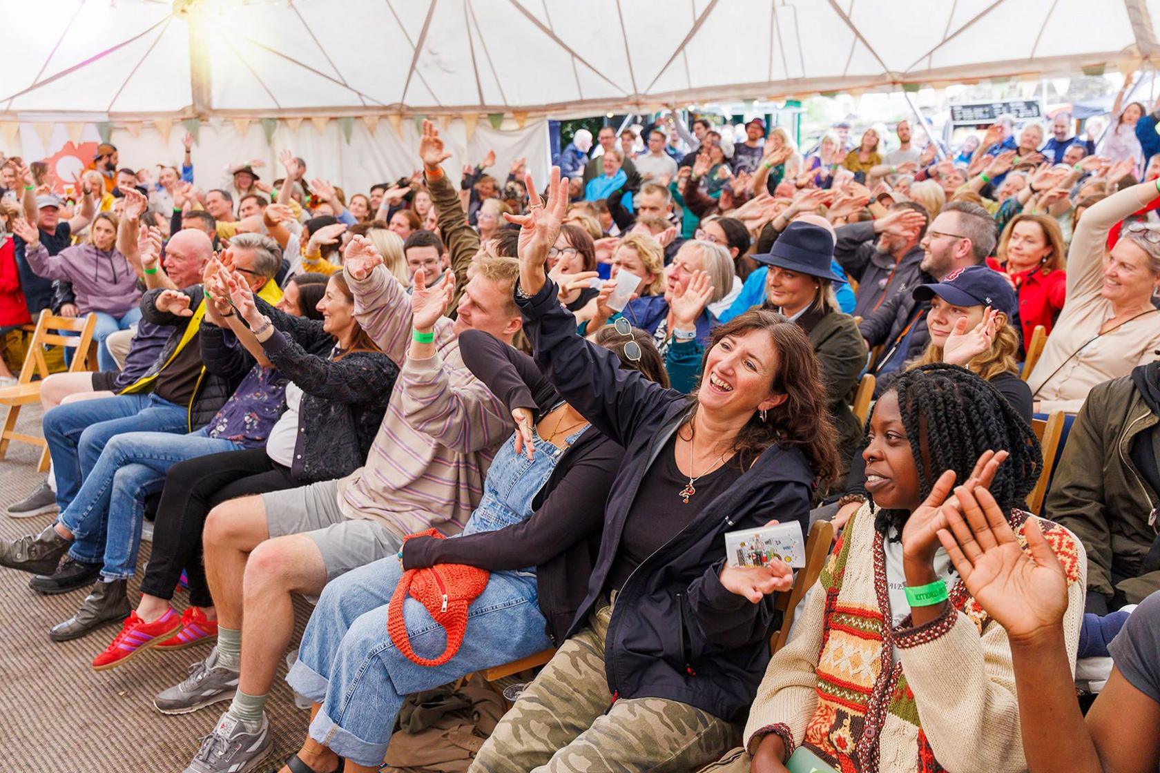 Lots of people in a well-lit large, white tent, siting on rows of seats. They are waving their arms back and forth as if they are taking part in some kind of crowd participation event, led by someone out of shot. Most are laughing and smiling.