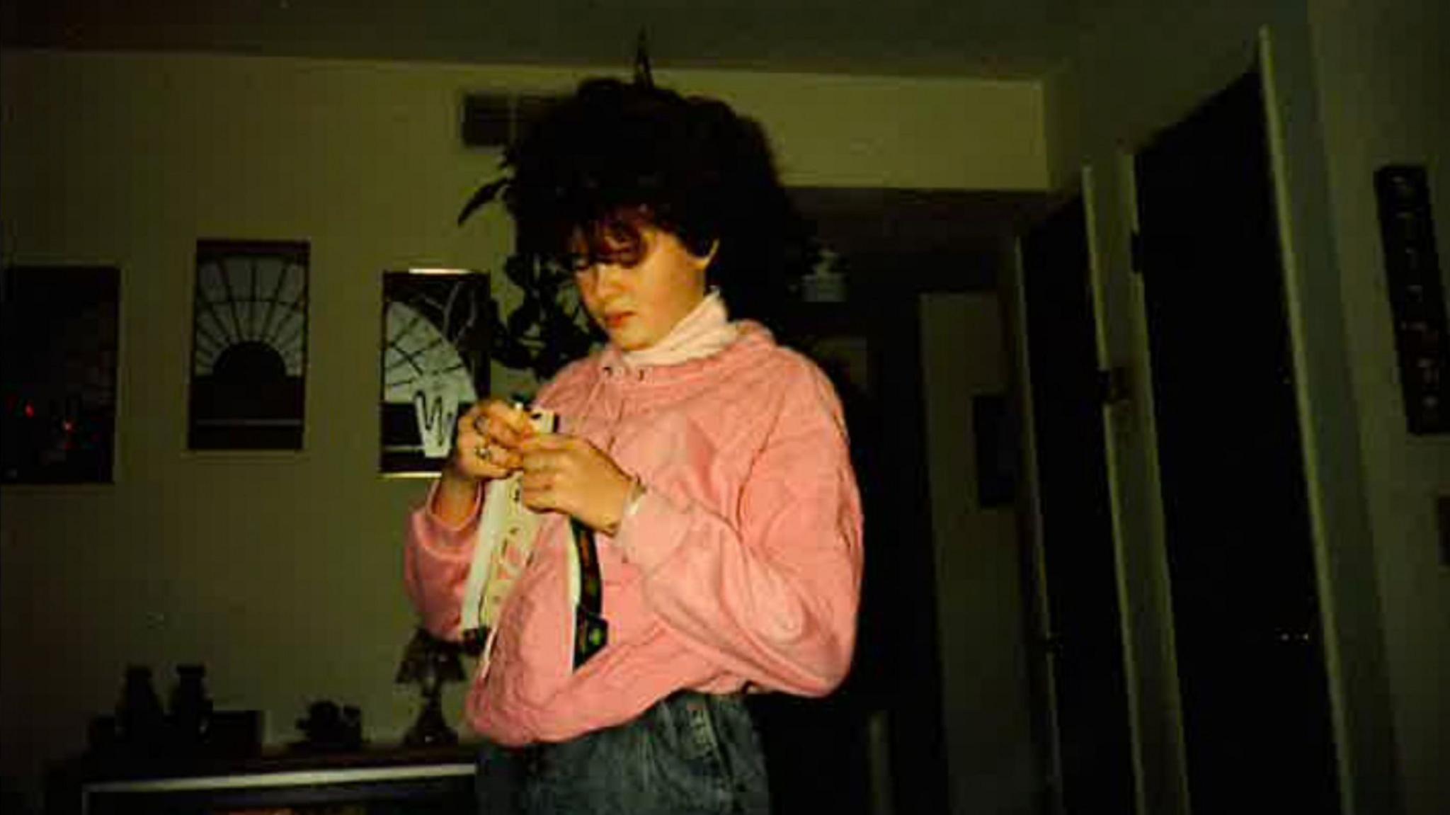 Christa Pike pictured as a teenager. She has curly brown hair and is wearing a pink top and jeans. She is looking down at something in her hands - the room is dark.