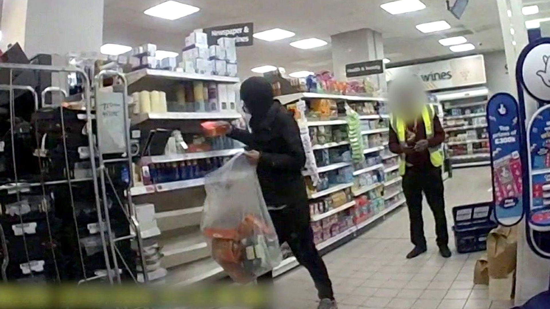 CCTV screenshot of a shoplifter in the act in a store aisle. The shoplifter is wearing a black hoodie and is in the process of filling a large transparent bag with goods, while a shopkeeper in high vis stands by.