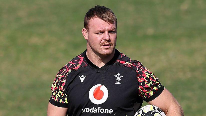 Dewi Lake wearing a black and patterned Wales branded training t-shirt holds a rugby ball under his left arm and looks straight ahead, as he stands on a pitch during a training session.
