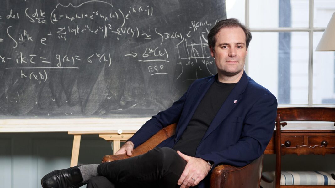 Reform receives record £36m donation from crypto billionaire Ben Delo sits in front of a blackboard in a brown leather armchair. He has short brown hair and wears a blue blazer, black t-shirt, black trousers, grey socks and black loafers. A maths equation is written on the board behind him.