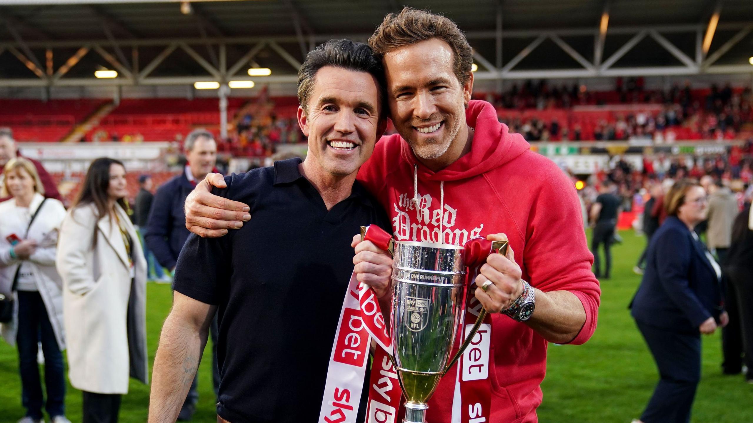 Ryan Reynolds and Rob Mac holding a trophy between them. They are both smiling and looking at the camera. Mac is wearing a black short sleeved polo shirt. Reynolds is wearing a red hoodie. They are stood on the pitch of The Racecourse Ground. People can be seen also on the pitch behind them.