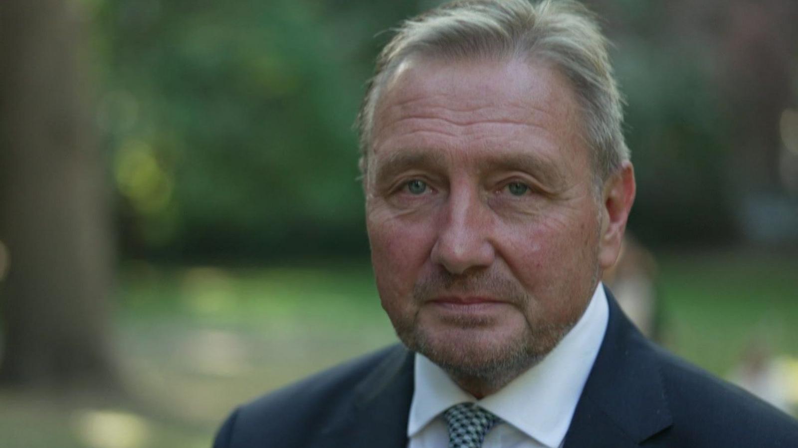 A man in late middle age wearing a dark blue suit, white shirt and patterned tie being interviewed in a park.