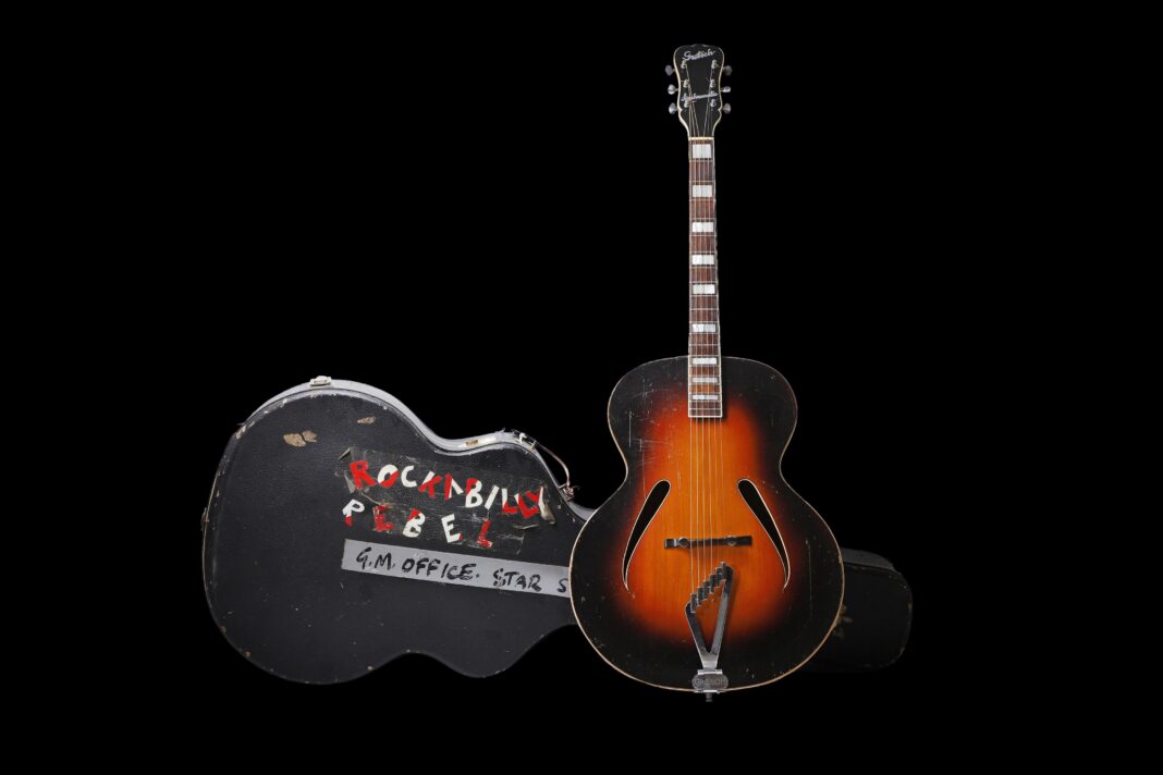 George Michael’s ‘Faith’ guitar up for auction An acoustic guitar with a sunburst pattern, standing next to a hard, black guitar case with the words Rockabilly Rebel written on it in red and white capital letters. The case and guitar are against a black background.