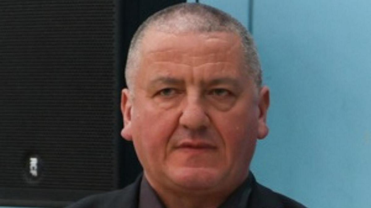 Andrew Harrison is wearing a black suit with a dark red tie with a light blue Reform rosette. He has close cropped grey hair.