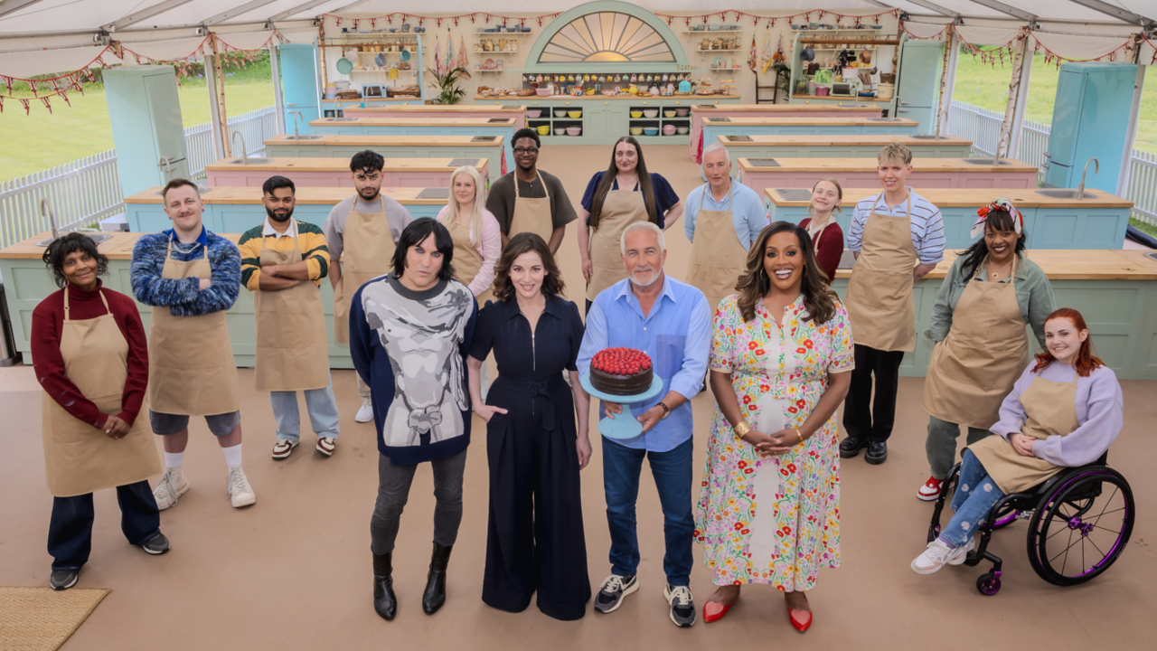 The 12 Bake Off contestants stand in a line behind the four presenters in the Bake Off kitchen tent