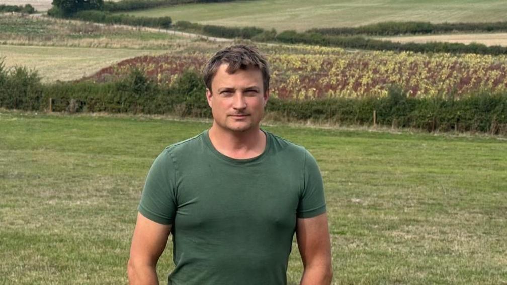 Farmer Tim Smith is pictured in a field. He is wearing a dark green T-shirt and has a serious expression on his face. There are further fields behind him.