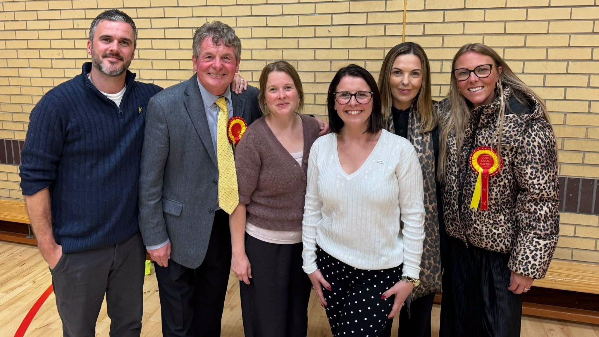 Sarah Maltby stands at the count and smiles, her team surround her and wear yellow and red badges.