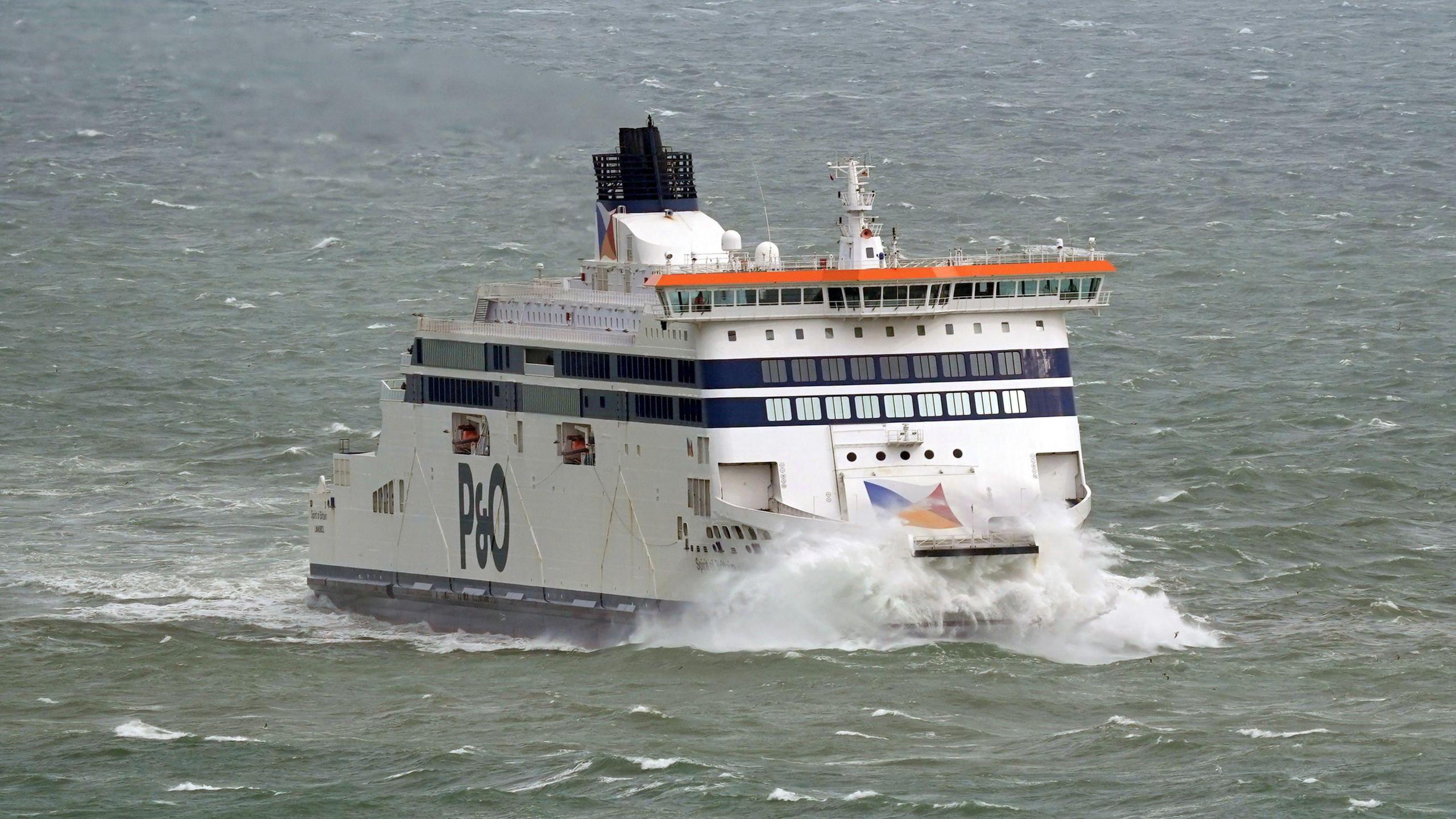 A P&O Ferries vessel travels through choppy sea conditions, with waves breaking against the bow. The white ferry has dark blue bands across the passenger decks and a large P&O logo on the side. The vessel is photographed from an elevated angle with open water surrounding it.