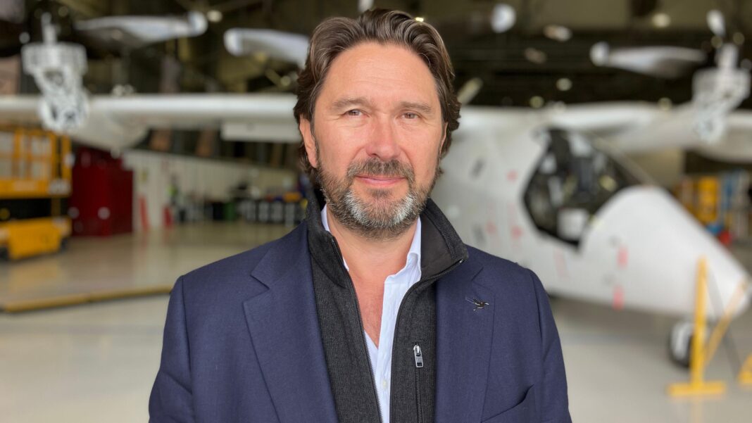 A man pictured in front of a small aircraft. He has short dark hair and facial hair and is wearing a blue blazer over a grey jacket. He is looking at the camera and smiling.