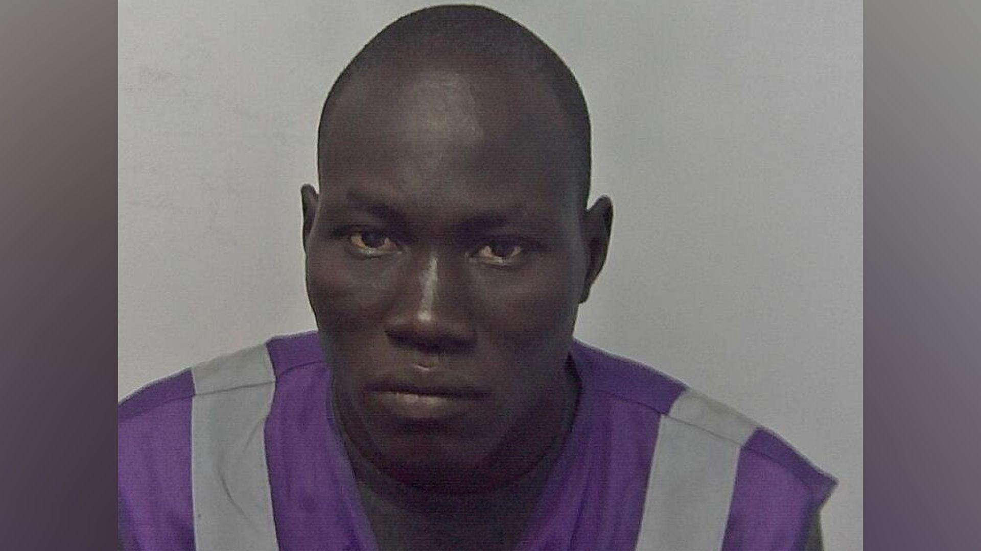 A mugshot of a man with a shaved head looking at the camera. He is wearing a purple and grey shirt.