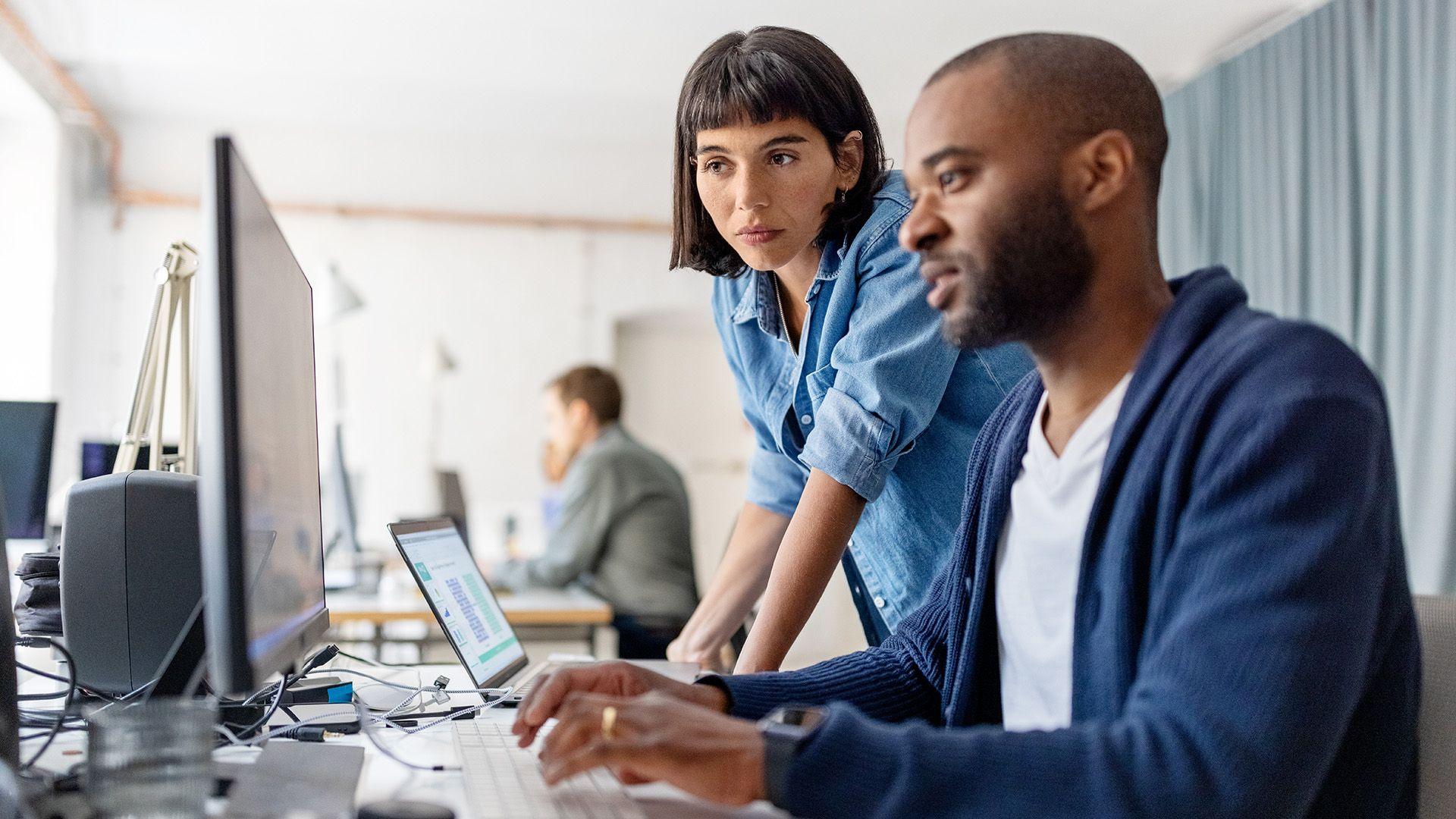 Stock photo shows a person leaning over the computer of another person in an office environment