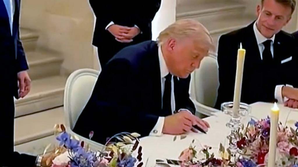 Donald Trump signing a document at a table with white table cloth and flowers. Macron is seen to his right watching