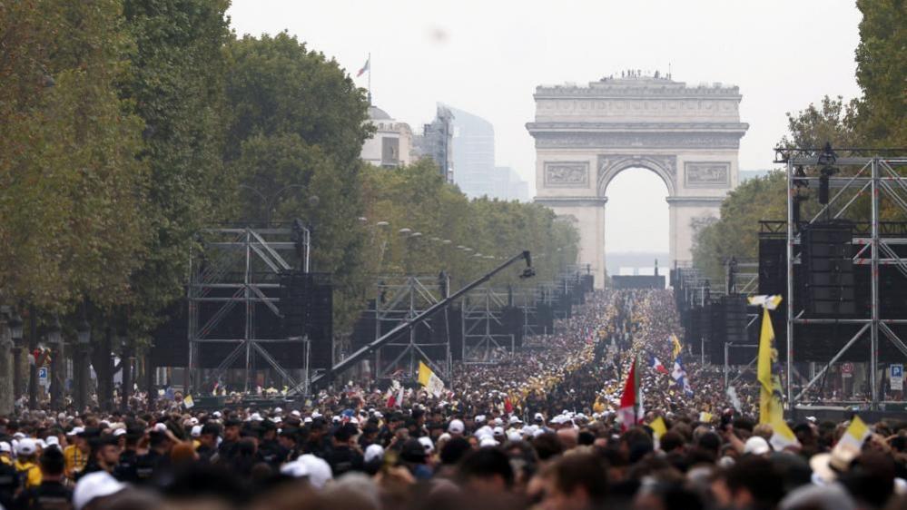 Thousands of people can be seen lining Champs-Elysees Avenue. A sea of white hats and yellow tops line the street. There are several large scaffold structures with screens on and the Arc de Triomphe can be seen at the end of the street.