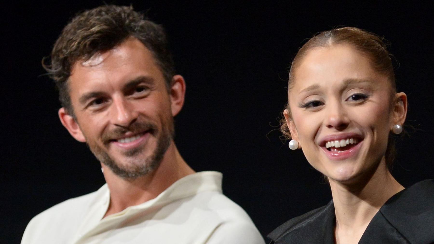 Jonathan Bailey, wearing a white open-necked shirt, and Ariana Grande in a black top and pearl stud earrings, at a Q&A on the film Wicked: For Good in Los Angeles. They are both laughing.