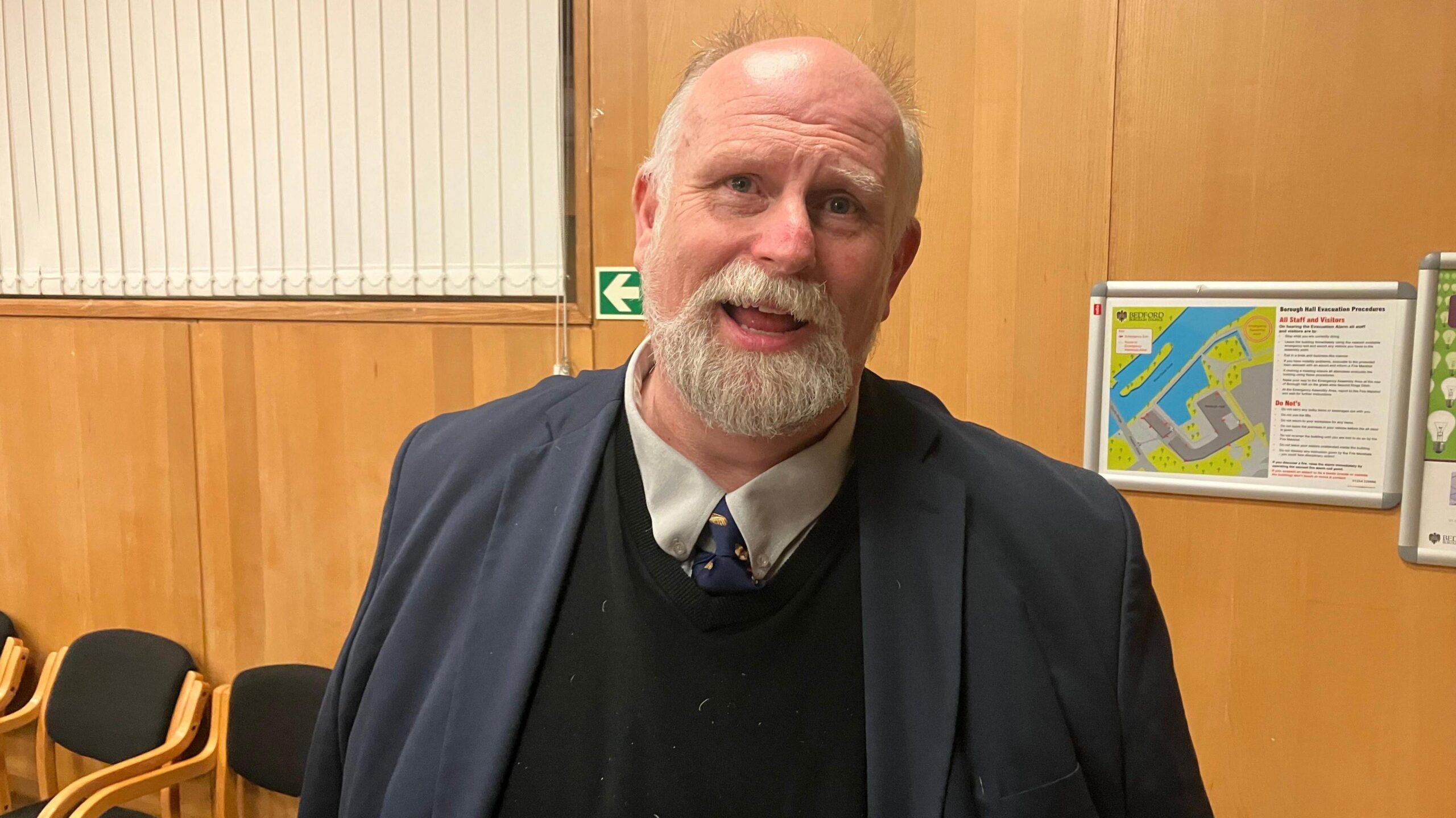Tom Wootton is looking into the camera while standing in a wood-panelled room that appears to be an office or meeting space. He has white hair on the sides of his head and a beard. He is dressed in a dark blue suit jacket over a black sweater, with a white collared shirt and patterned tie. Behind him are wall-mounted evacuation diagrams and a row of black cushioned chairs with wooden frames.