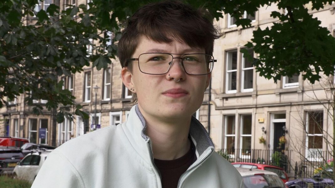 ‘My rent was higher than my student loan’ – the booming cost of private halls Lara wearing glasses and a light-coloured zip-up fleece standing outdoors beneath leafy trees, with stone terraced buildings and parked cars visible in the background.