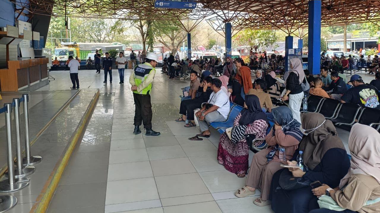 People sit and wait inside a busy ferry terminal, while a security guard stands nearby beneath directional signs and an open metal roof