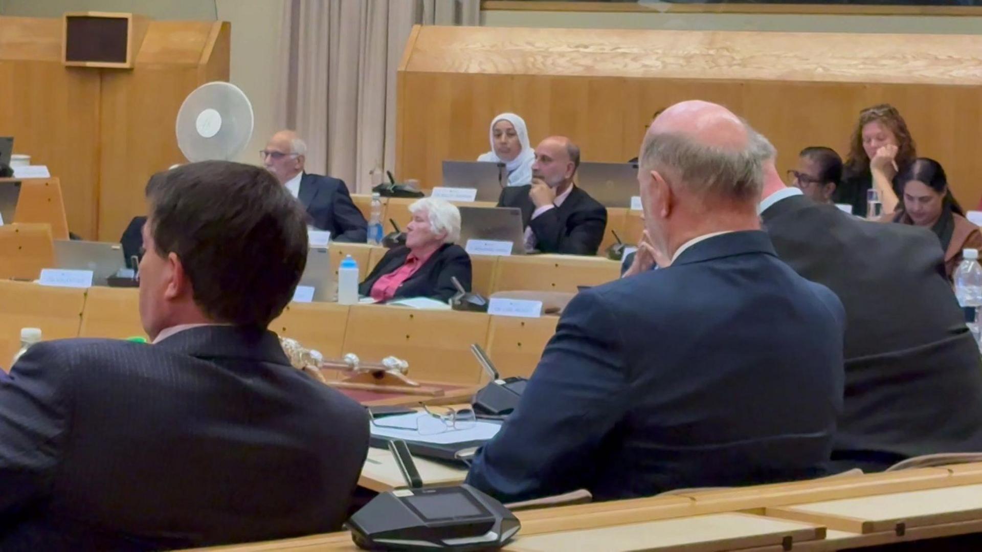 Members of Bedford Borough Council sit in a modern council chamber during a meeting. Several people are seated at curved wooden desks equipped with microphones, laptops and documents. In the foreground, attendees in dark suits are seen from behind, while others sit and listen across the room. A ceremonial mace rests on a desk near the centre of the chamber. Light wood panelling, a large fan and computer screens are visible in the background.