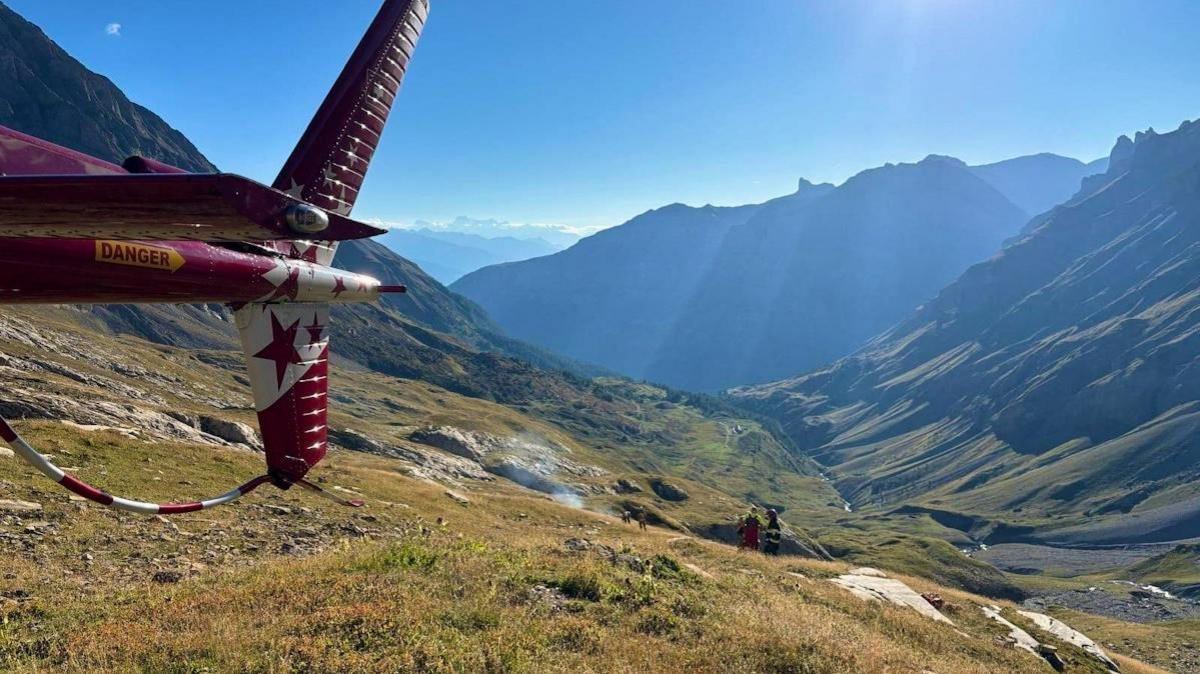 Helicopter tail in foreground with rising smoke in the distance and emergency crews seen high in the Alps overlooking a mountain range
