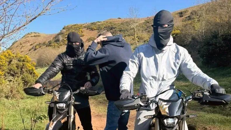 Two young men with black masks on riding electric motorbikes on a path in a rural area